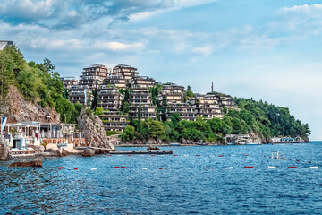 Naklejka premium A scenic coastal view of Dukley Gardens in Budva, Montenegro, with luxury residential buildings perched on a hillside overlooking the Adriatic Sea. The waterside resort area with a restaurant