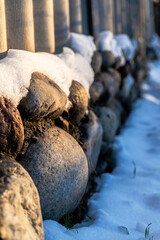 A detailed close-up of a rustic stone wall partially covered with snow. The sunlight enhances the natural textures of the smooth stones and melting snow, creating a serene winter atmosphere. 