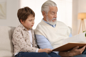 Grandpa and his grandson reading book together on sofa at home