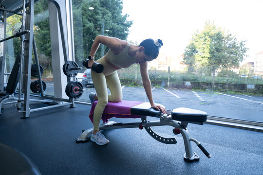 Female athlete performing one arm dumbbell row, kneeling on weight bench in bright gym, demonstrating focused strength training technique and muscular fitness preparation