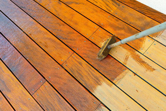 Close-up of a wooden deck being stained with a brush for protection and maintenance during a home improvement project in bright sunlight