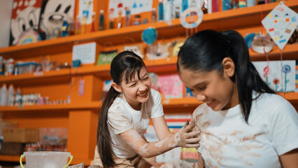 Happy multicultural girl and friend put the clay on the each other muddy shirt at art lesson. Diverse highschool student playing with clay at pottery workshop. Creative activity concept. Edification.