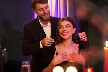 Affectionate man presenting luxury necklace to his surprised girlfriend at romantic dinner in restaurant. Valentine's day celebration