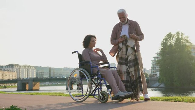 Senior woman in wheelchair walking with caregiver old man on road. Elderly family couple man supporting taking care of paralyzed woman in chair for people with disability outdoor. Rehabilitation