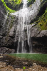 The breathtaking view of a stunning cascading Madakaripura Waterfall flows gracefully into a tranquil green pool, beautifully surrounded by mossy rocks and vibrant nature all around, Java, Indonesia