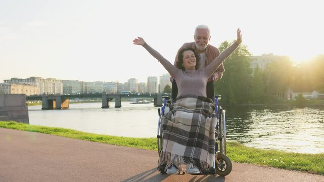 Senior woman in wheelchair walking with caregiver old man on road. Family couple man husband supporting taking care of paralyzed woman wife in chair for people with disability outdoor. Rehabilitation