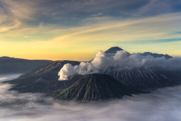 A breathtaking view of a Bromo, volcanic region at sunrise, featuring soft clouds gracefully rolling over jagged peaks and mist gently enveloping the serene valleys below, East Java, Indonesia