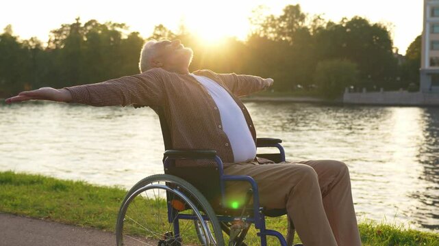 Happy senior man in wheelchair raised hands enjoying freedom walking on road in city park outdoor. Old happy man in chair for people with disability has a hope. Disability disease rehabilitation