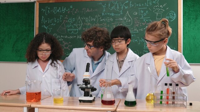 Smart caucasian teacher looking under microscope while diverse children doing experiment at STEM science lesson. Professional happy scientist inspect biochemical example by using microscope. Pedagogy.