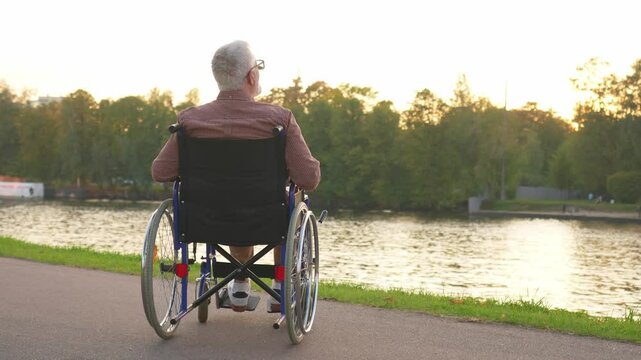 Sad senior man sitting in wheelchair walking on road in city park outdoor, back view. Old paralyzed man in chair for people with disability feel depressed. Lonely elderly sick man sits on wheelchair