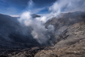 A breathtaking volcanic crater, Bromo volcano, is actively emitting smoke, surrounded by majestic towering mountains that rise high beneath a clear blue sky, creating a stunning view, Java, Indonesia