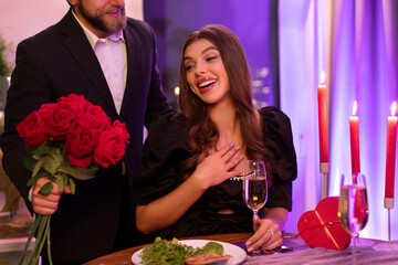 Man in suit giving red roses to his happy girlfriend, couple having romantic date at restaurant in the evening, celebrating Valentine's day