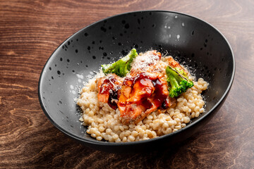 A delicious dish featuring grilled salmon, broccoli, and a savory sauce served over pearl couscous. Presented in a stylish black bowl on a wooden table, perfect for gourmet menus.