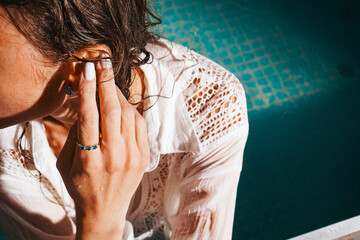 Stylish woman leaning on the pool edge, wearing a white blouse with wet hair and delicate jewelry.