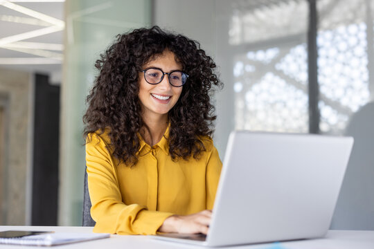 Joyful and satisfied woman working with laptop inside office, businesswoman typing on keyboard while sitting at desk, office worker smiling wearing shirt and glasses. - Powered by Adobe