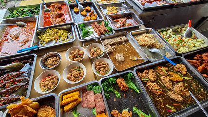 Vibrant display of various cooked dishes at food stall or buffet. Dishes are in metal trays and bowls, showcasing a variety of meats, vegetables, and sauces. Street food in Southeast Asia