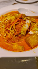 Close-up of a plate of curry mee or a similar curry noodle dish. Yellow noodles are immersed in a creamy, orange-red curry broth with tofu puffs and possibly other ingredients like vegetables