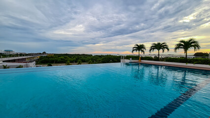 Naklejka premium Infinity pool with calm, blue water reflects cloudy sky. Palm trees near pool's edge, overlooking coastal landscape with greenery and buildings in distance. Tranquility and luxurious vacation setting