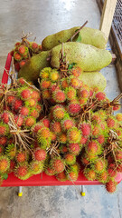 Fototapeta premium Collection of rambutan and jackfruit are displayed on a red surface of cart. Bunches of rambutan, with their characteristic red and green spiky exteriors. Exotic fruit market in Melaka, Southeast Asia