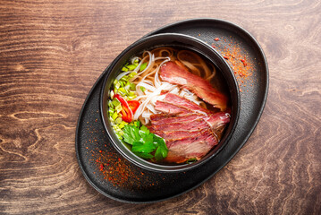 A close-up of a flavorful beef noodle soup served in a black bowl. The dish features tender slices of beef, fresh herbs, and red chili, all presented on a wooden table.