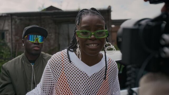 Chest up handheld shot of young African American lady in glasses and classy outfit rapping to camera while filming music video in shabby area, cool guy singing in background, cameraman recording