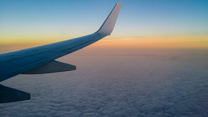 Fototapeta premium Plane's wing extends into a vast expanse of fluffy white clouds, seen from an airplane window. Sky above the clouds transitions from pale blue to warm orange-yellow near horizon, sunrise or sunset