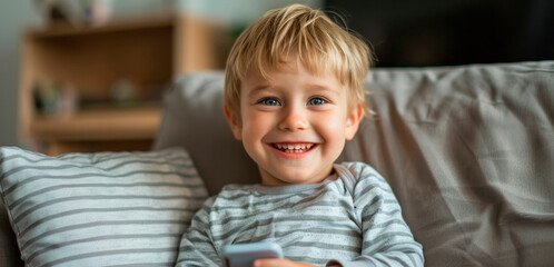 Fototapeta premium A cheerful baby grins widely while holding a phone, sitting comfortably on a couch with striped cushions. The cozy living room is softly lit, creating a warm atmosphere for play.