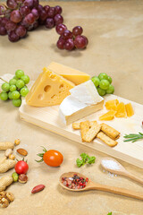 Several types of cheese on a light wooden board with fruits, vegetables and herbs