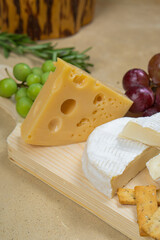 Several types of cheese on a light wooden board with fruits, vegetables and herbs