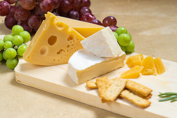 Several types of cheese on a light wooden board with fruits, vegetables and herbs