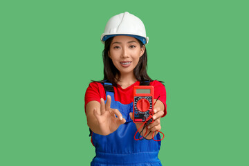 Young female electrician with multimeter showing OK gesture on green background