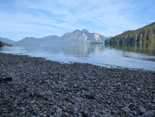Kiesstrand an einem ruhigen See mit Bergspiegelung und klarem Himmel.
Pebble beach by a calm lake...