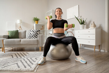 Sporty young woman with dumbbells training on fitball at home