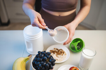Athletic woman in sportswear preparing protein drink in a kitchen, putting a portion of whey protein powder from a measuring spoon into a shaker in her hand