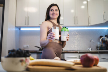 Attractive, smiling woman in sportswear holding a cocktail shaker and a glass with a protein drink,...