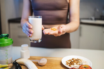 Athletic woman in sportswear holding a glass with a protein drink, milkshake, or smoothie and omega-3 capsules, fish oil supplement in her hand, in a kitchen
