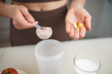 Athletic woman in sportswear preparing protein drink in a kitchen, holding a measuring spoon with whey protein powder and omega-3 supplements, white amino acid capsules or vitamins in her hand