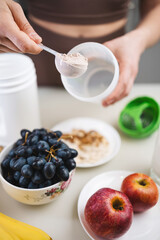 Athletic woman in sportswear preparing protein drink in a kitchen, putting a portion of whey protein powder from a measuring spoon into a shaker in her hand