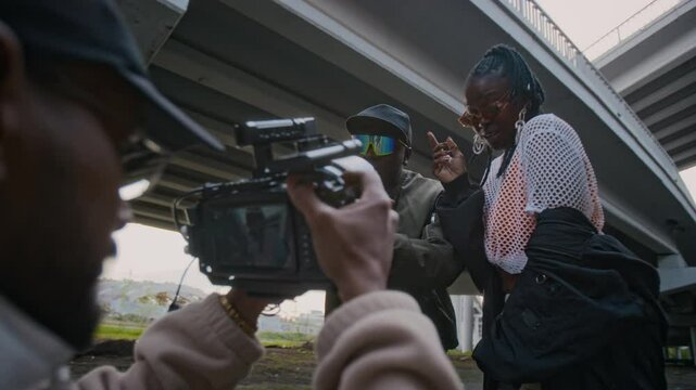 Over shoulder shot of male videographer shooting two African American female and male artists rapping and vibing to hip-hop music for camera, wearing stylish outfits, recording music video outdoors