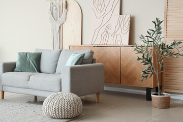 Interior of light living room with grey sofa, olive tree and chest of drawers