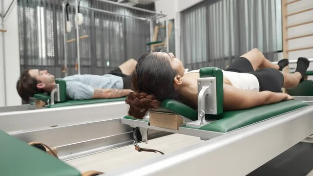 Couple practicing leg exercises on pilates reformer to strengthen core and improve flexibility at gym. Pilates partner doing pelvic bridge pose on reformer for improving lower back muscle. Habituate.