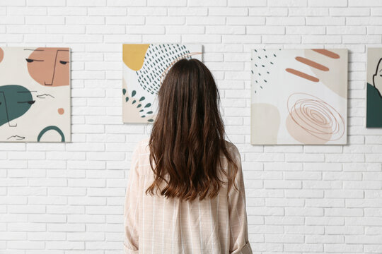 Young woman near paintings on white brick wall at art gallery exhibition, back view