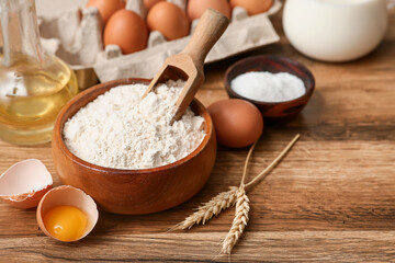 Bowl of flour with scoop, eggs, wheat spikelets and salt for preparing dough on wooden background