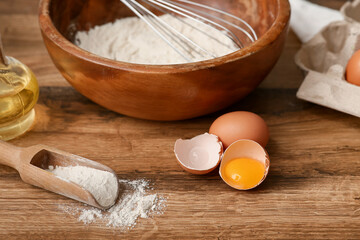 Scoop with flour, bowl with whisk and eggs for preparing dough on wooden background
