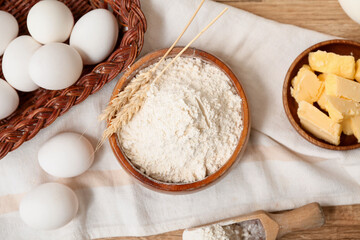 Bowl of flour with scoop, eggs and butter for preparing dough on wooden background