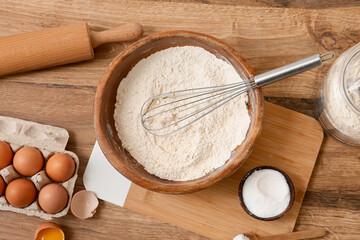 Bowl of flour with whisk, eggs, salt and rolling pin for preparing dough on wooden background