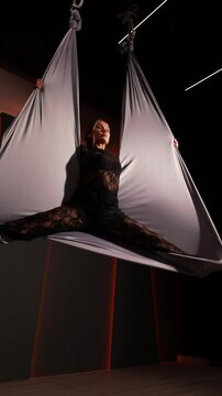 Aerial dancer performing acrobatic stretches inside suspended gray hammock, wearing black outfit with dramatic dark background and red lighting