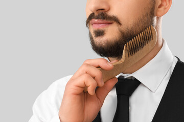 Handsome young bearded man combing beard on grey background, closeup
