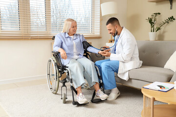 Fototapeta premium Male doctor measuring blood pressure of mature woman in wheelchair at home