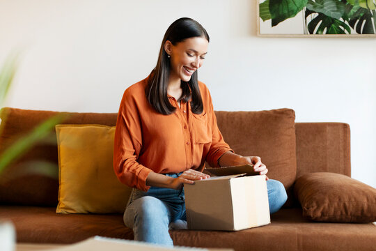 My online order is here. Happy young woman receiving package, sitting on sofa and opening carboard box at her home interior